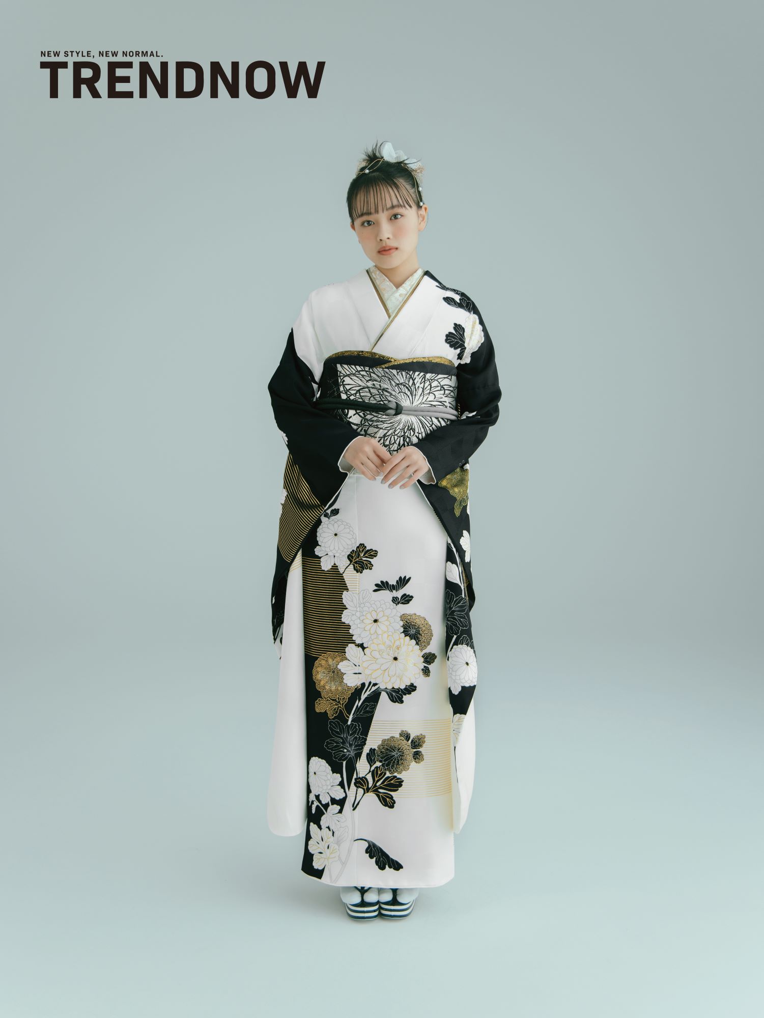 黒、白、金の花柄が入った日本の伝統的な着物を着た人物が立っています。白い花の髪飾りをしており、明るい灰色の背景にポーズをとっています。左上隅に「TRENDNOW」という文字が表示されています。.