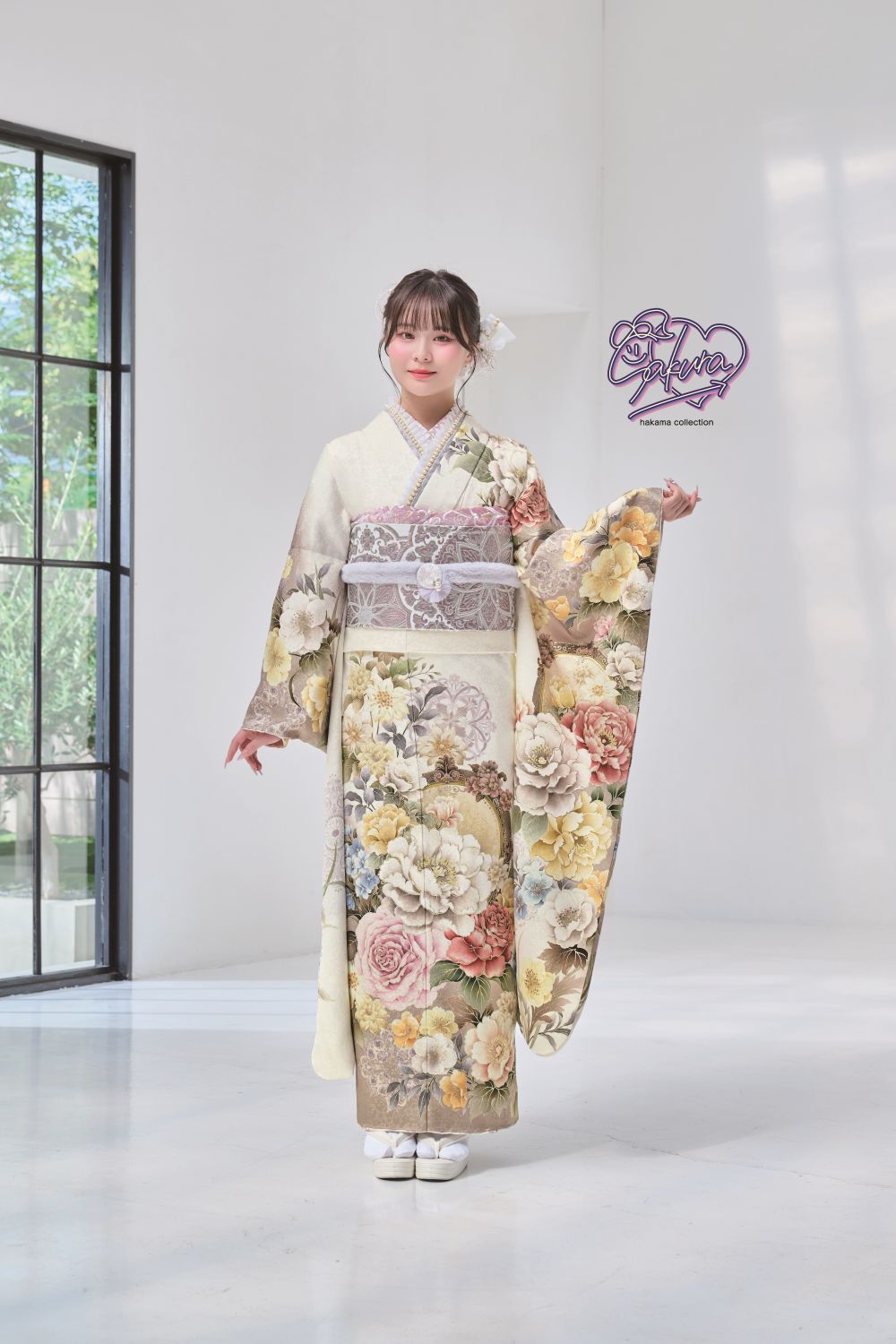 ベージュ、ピンク、黄色、緑の色合いの花柄が描かれた装飾的な着物を着た人が室内に立っています。髪には白い花のアクセサリーをつけています。部屋には白い壁と、黒い枠の大きな窓があり、左側から日光が差し込んでいます。.