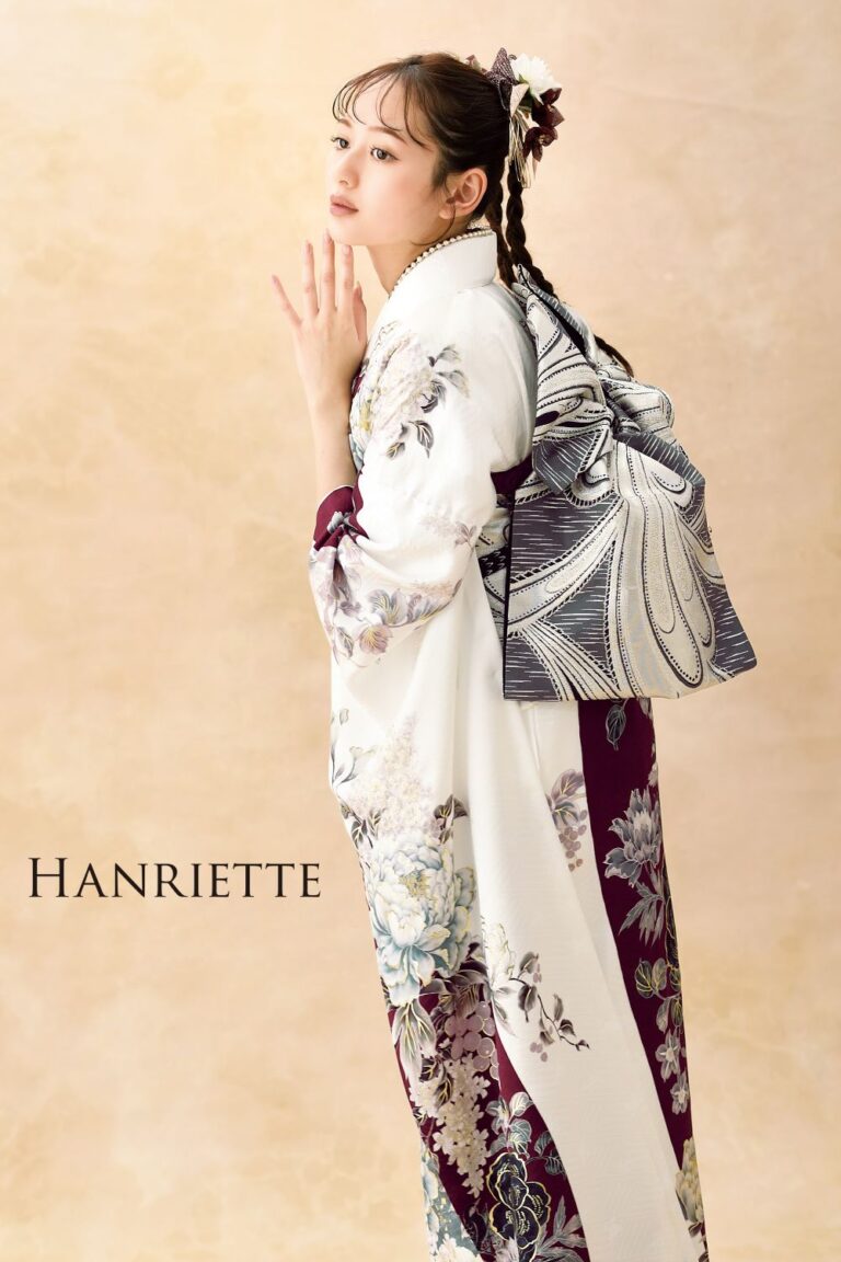 女性が、白と紫の花柄の着物を着て、後ろで複雑な白と黒の帯を結んで優雅に立っています。彼女の髪は三つ編みでまとめられ、花で飾られています。彼女は柔らかいベージュ色の背景を背に横向きでポーズをとっています。画像には「HANRIETTE」という文字が表示されています。.