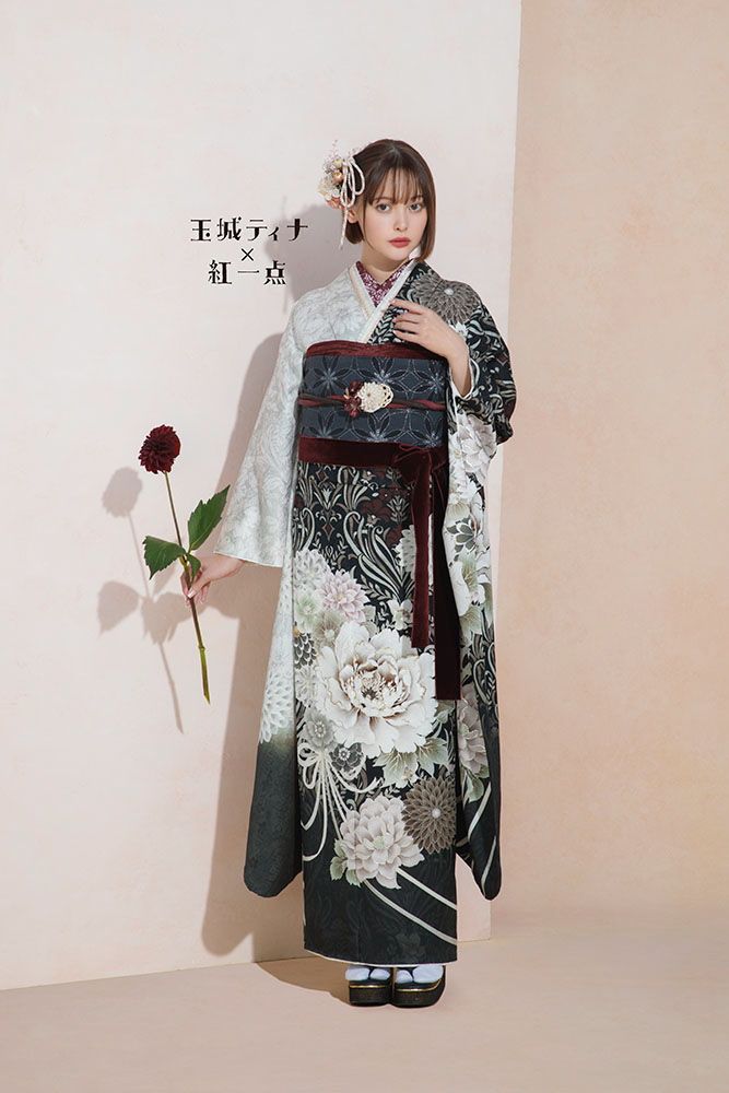 女性が、白とグレーの複雑な花柄で飾られた伝統的な日本の着物を着て立っています。彼女は濃い赤の花を一輪持っています。彼女の髪は、それに合わせた花のアクセサリーで飾られています。背景は、シンプルな黄褐色と白の壁です。彼女の横には日本語の文字があります。.