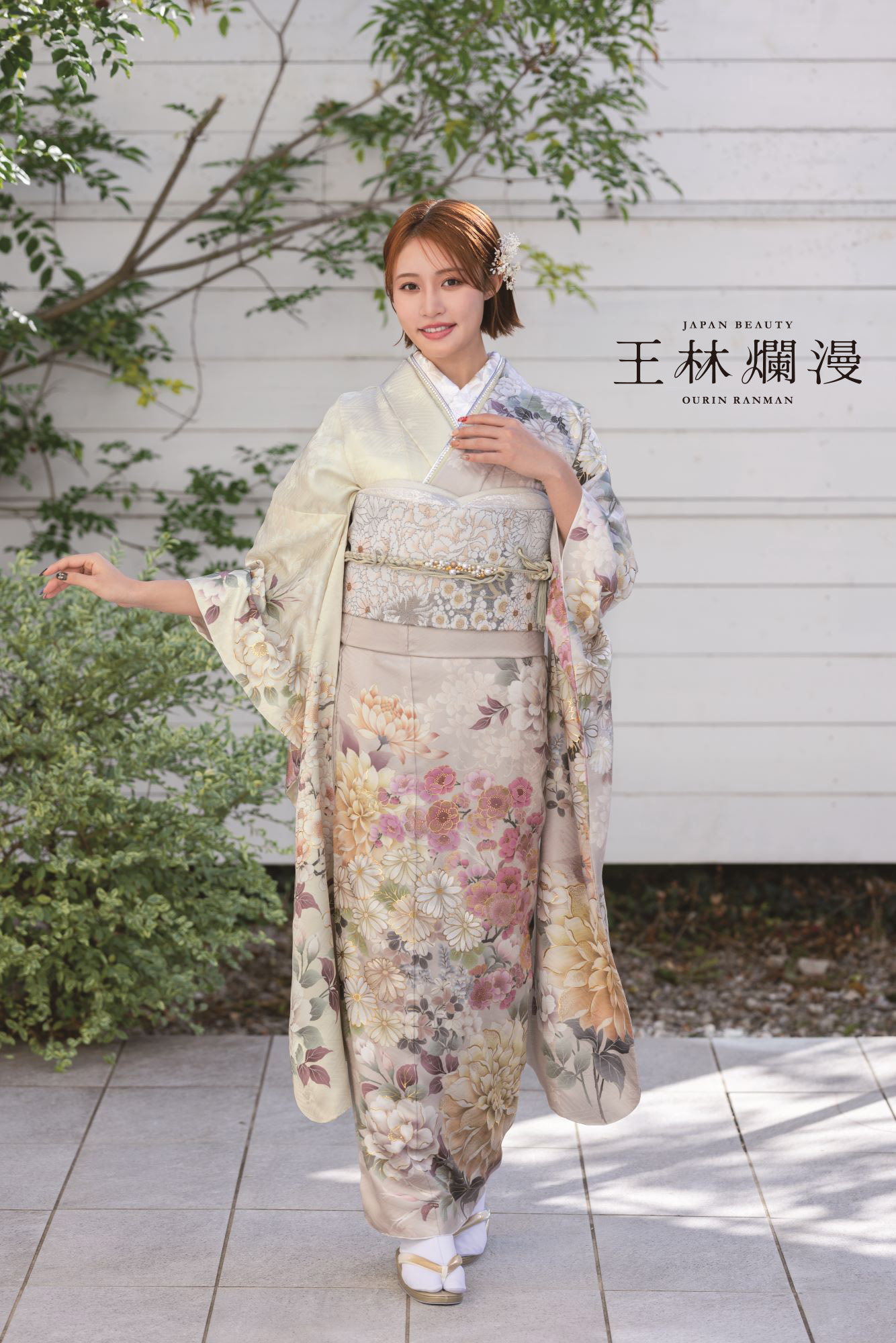 伝統的な花柄の着物を着た女性が、緑の木々に囲まれた白い木の壁の前の石畳の道に立っています。彼女は髪をエレガントにまとめ、片手を軽く上げて微笑んでいます。壁には「日本の美」と「桜林爛漫」の文字が書かれています。.
