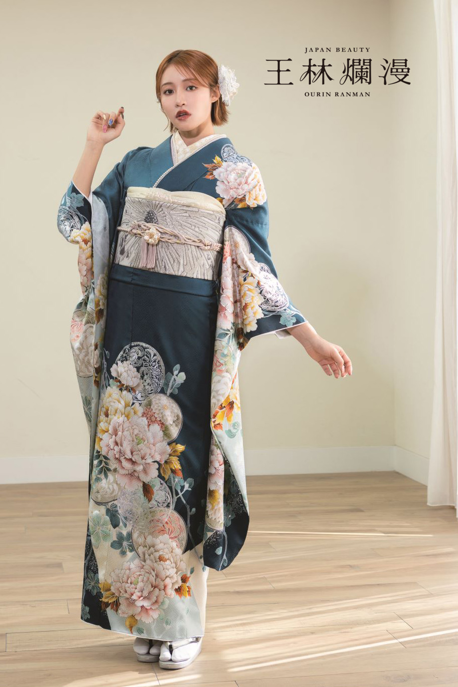 花柄の青緑色の着物を着た女性が、室内の木の床の上に立っている。着物にはピンクと白の牡丹が描かれ、腰にはクリーム色の帯が結ばれている。彼女は片手を顔の近くに、もう片方の手を体の横に置いてポーズをとっている。壁には日本語と英語の文字が見える。.
