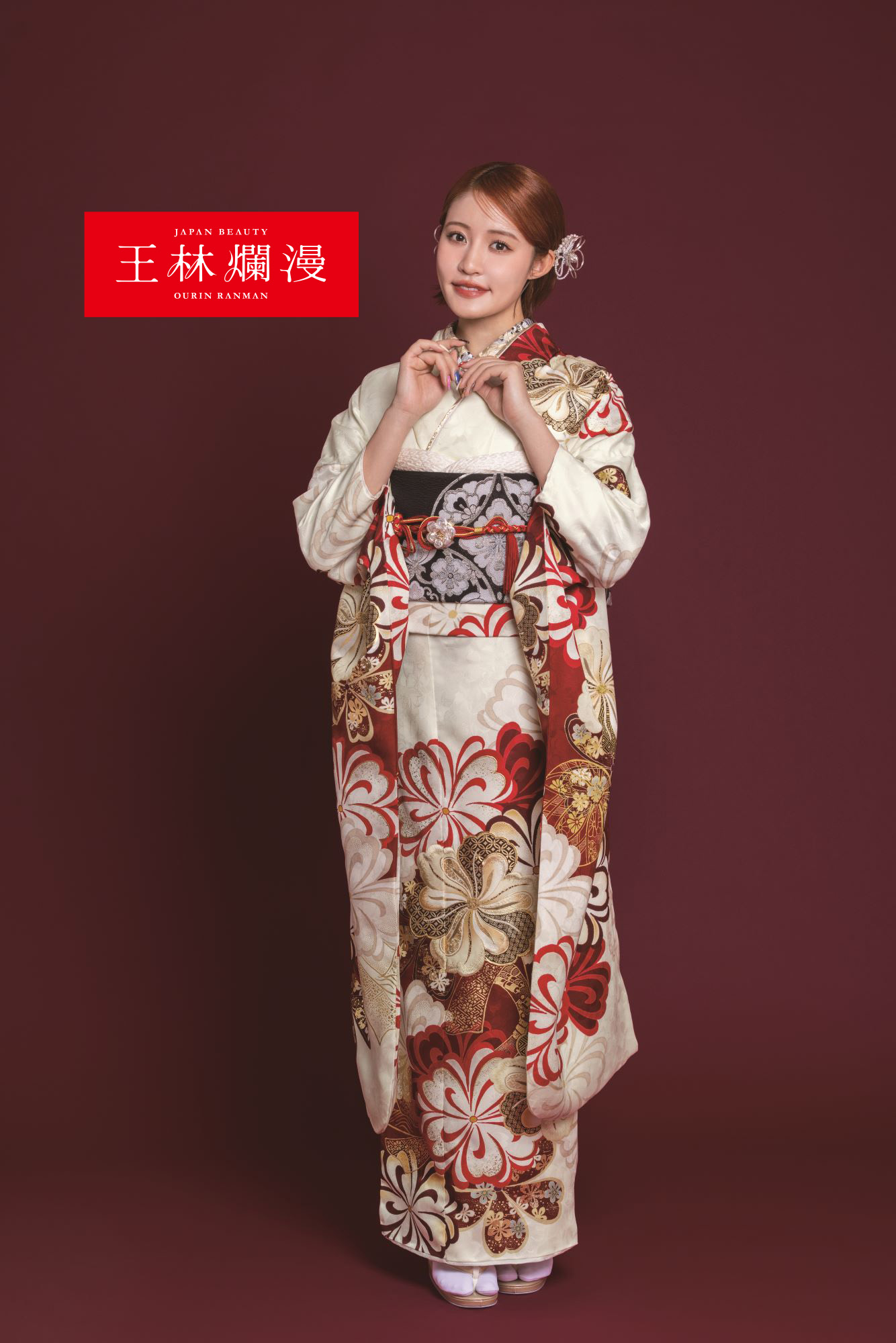 栗色の背景に、クリーム色と赤色の伝統的な花柄の着物と黒帯を身に着けた女性が立っています。彼女の髪は花飾りで飾られています。彼女は小さな物を持って微笑んでいます。左上隅には、白い文字が書かれた赤い長方形のラベルがあります。.