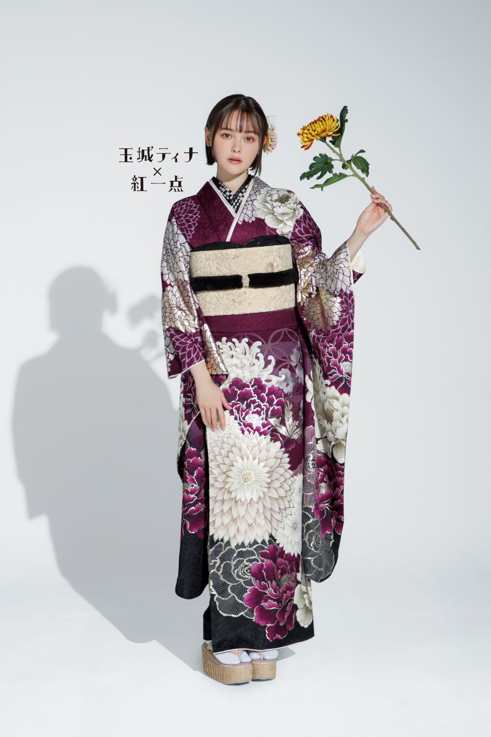 濃い紫と白の花柄が特徴的な日本の伝統的な着物を着た女性が立っています。右手には黄色い菊の花が一輪あります。背景は薄い灰色で、肩の近くに日本語の文字が見えます。彼女の視線はカメラに向けられています。.