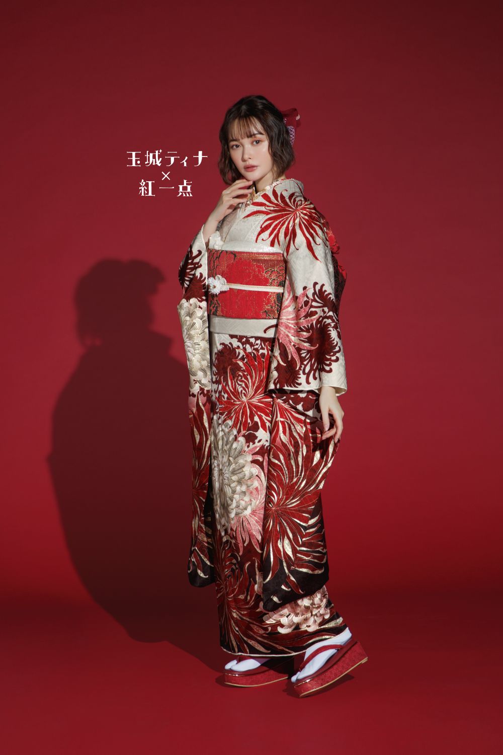 濃い赤の背景に、大胆な赤、銀、黒の花柄で飾られた豪華な着物を着た女性が立っています。彼女の髪はエレガントにスタイリングされており、伝統的な日本の草履を履いています。画像の左側には白い文字があります。.