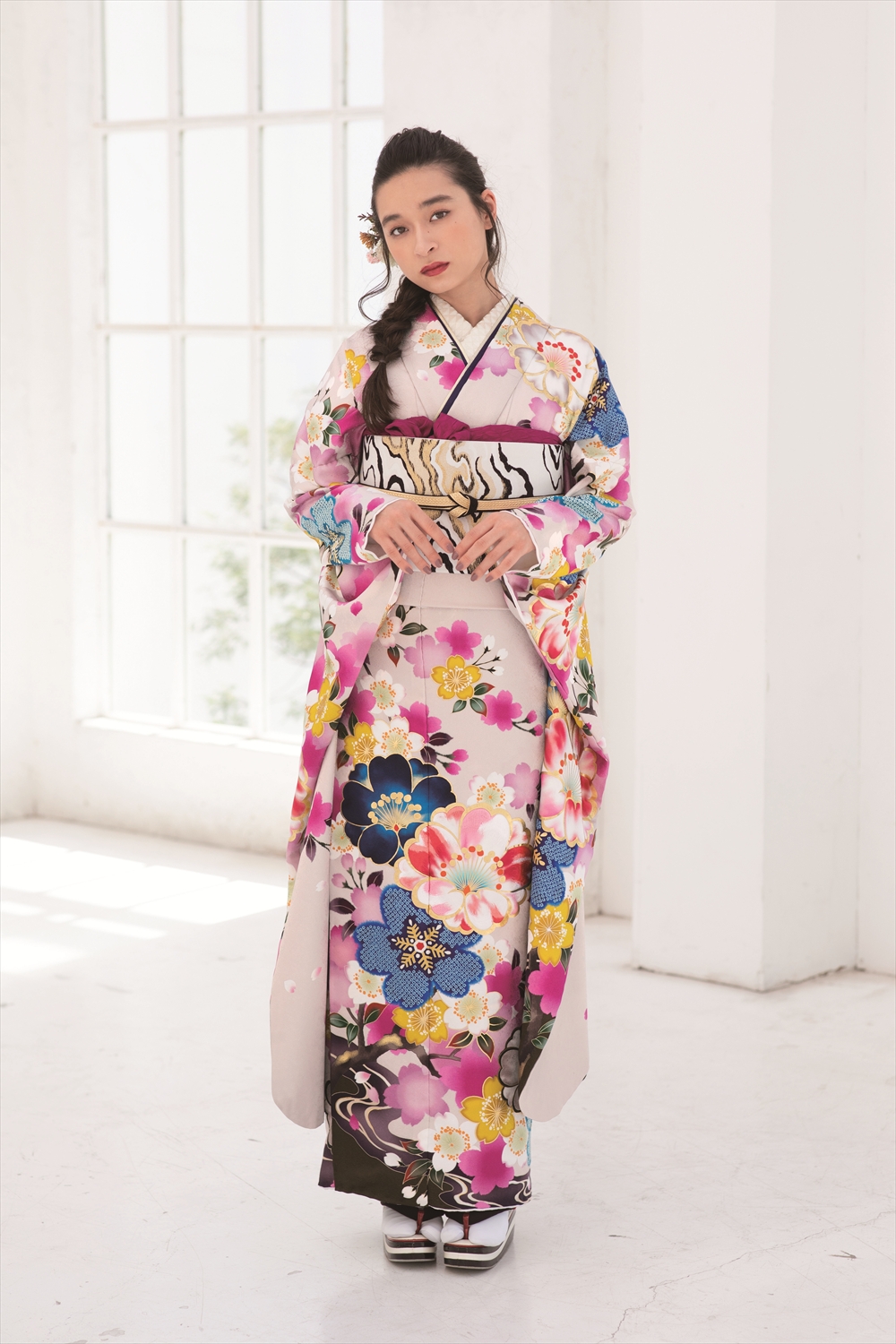 明るい部屋に、ピンク、青、黄色の花柄があしらわれた色鮮やかな着物を着た女性が立っている。長い黒髪をヘアアクセサリーで少し束ね、少し横を向いている。背景には白い壁と大きな窓が見える。.