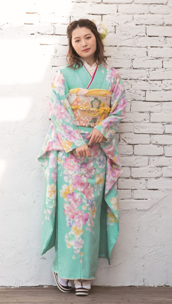 パステルグリーンにピンクと黄色の花が描かれた花柄の着物を着た若い女性が、白いレンガの壁を背に立っている。髪には花を飾り、白い靴下と草履を履いている。落ち着いた表情で、カメラをまっすぐに見つめている。.