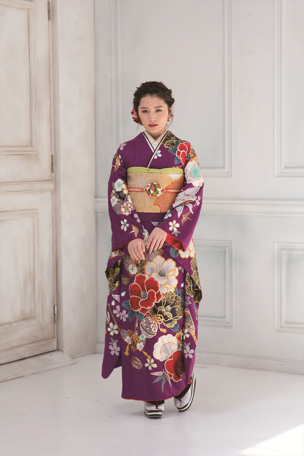 若い女性が、淡い色の壁を背に、鮮やかな紫色の着物に金色の帯を締めて立っている。髪には花飾りが飾られ、落ち着いた表情で両手を前に組んでいる。.