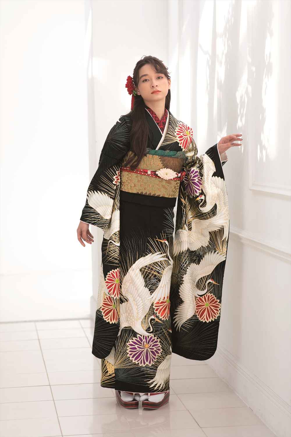 白い鶴と色とりどりの花が飾られた伝統的な黒い着物を着た女性が、室内に立っている。長い黒髪に赤い花の髪飾り、白い足袋に木靴を履いている。窓から差し込む光が、壁に柔らかな影を落としている。.