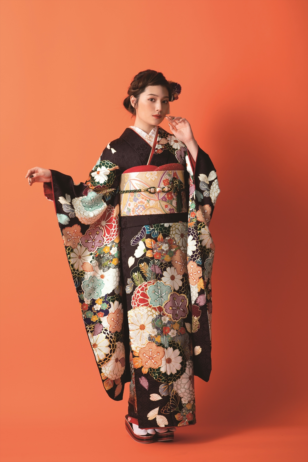 オレンジ色の背景に、色鮮やかな花柄があしらわれた黒い着物を着た若い女性が立っています。髪には花を添え、白い足袋と草履を履き、片手を顔に添え、もう片方の手で着物の袖を押さえ、優雅なポーズをとっています。.