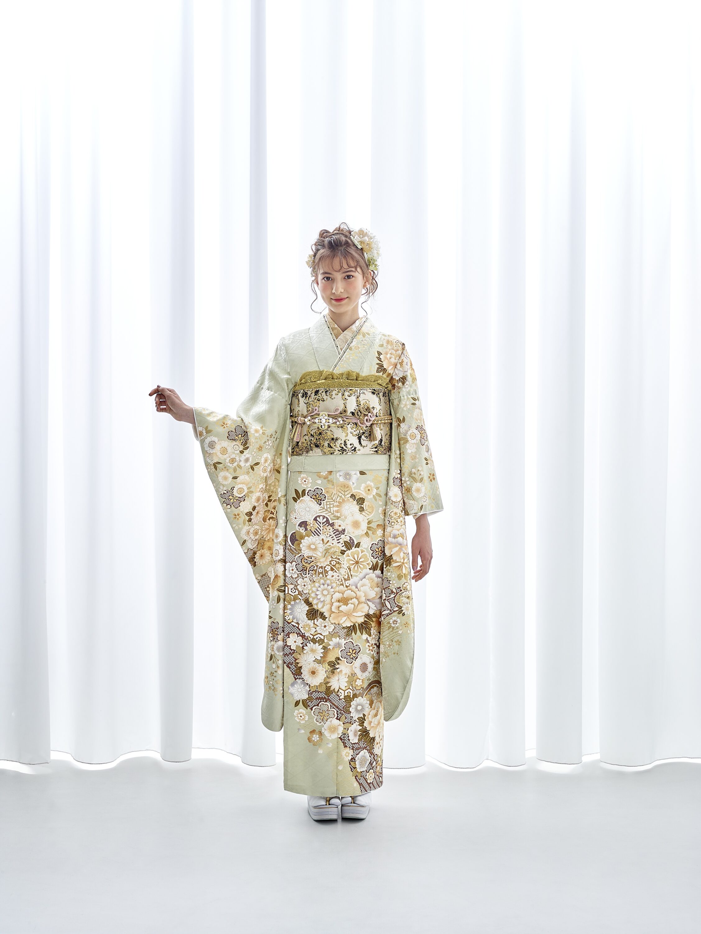 白いカーテンの前に、金、白、茶色の花柄があしらわれた淡い緑色の着物を着た女性が立っている。髪には飾りの簪が飾られ、白い草履に白い足袋を履いている。.