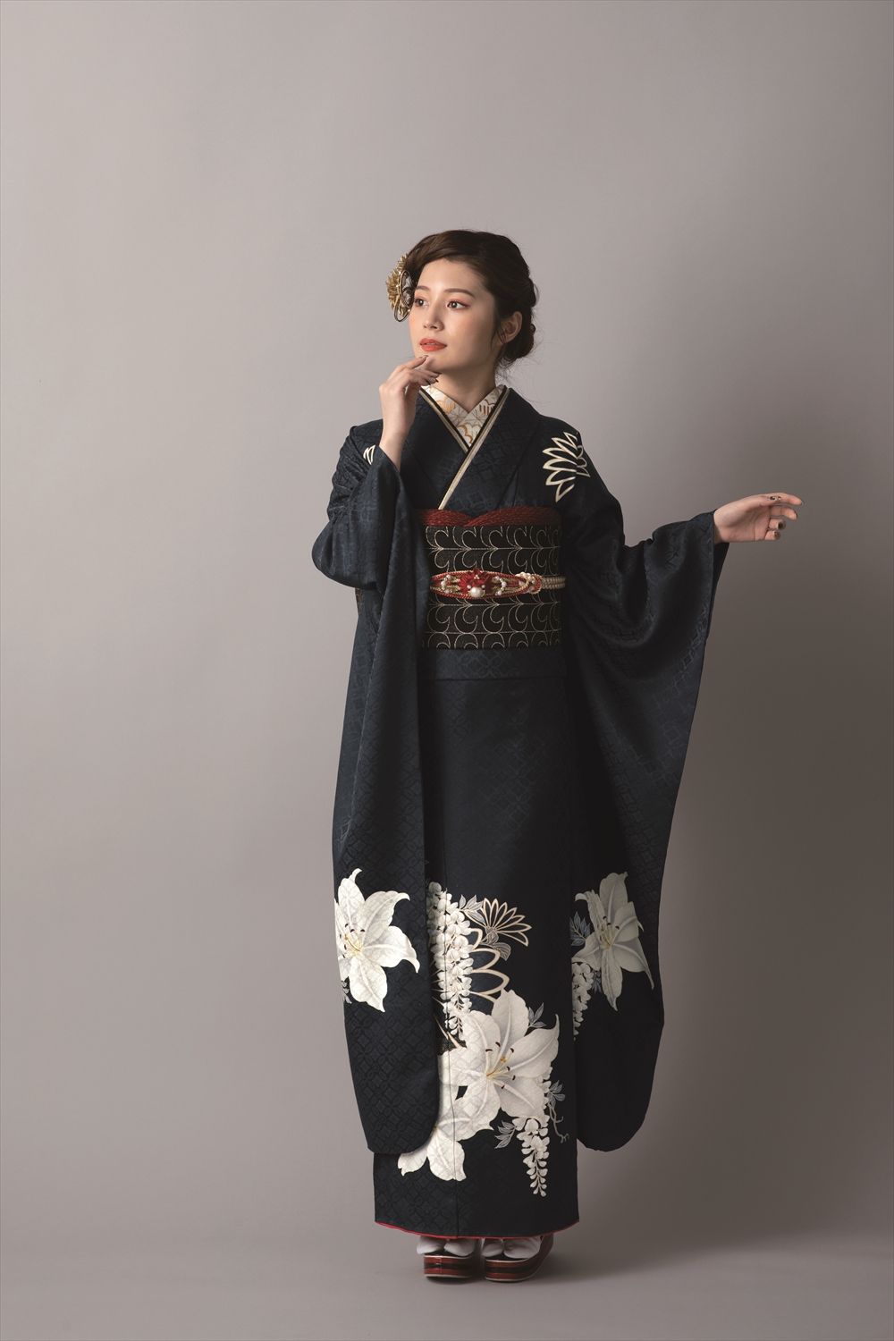 大きな白い花柄があしらわれた黒い着物を着た女性が、ニュートラルな背景を背に立っている。髪は飾りの簪できちんとセットされ、片手を挙げて着物の長い袖を際立たせながら、横を向いて優雅なポーズをとっている。.