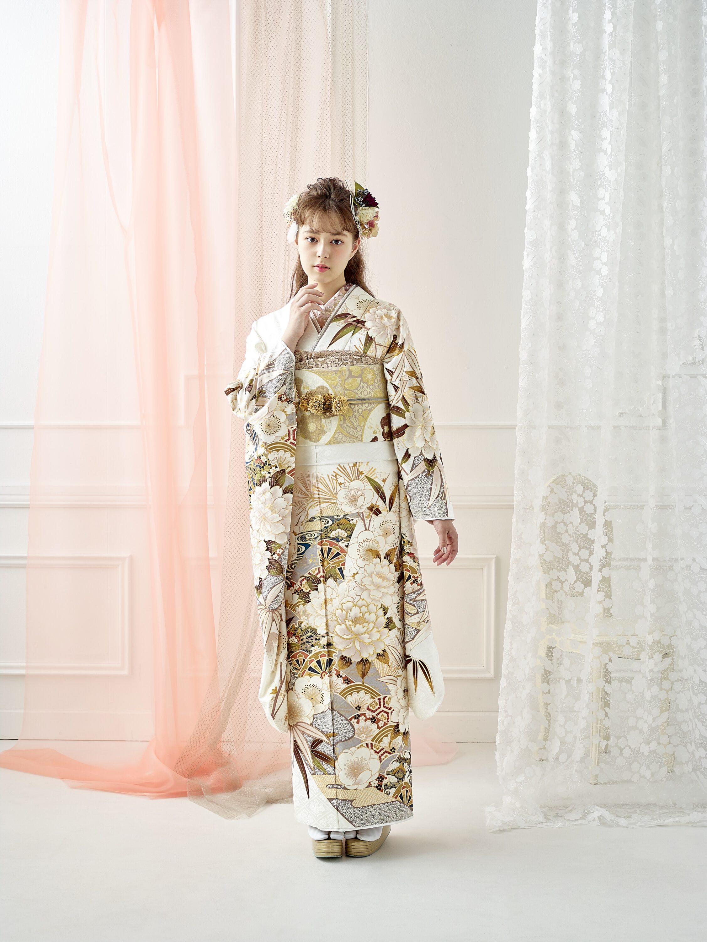 白と金の花柄の着物に華やかな帯を締めた若い女性が、室内に立っています。髪には花を飾り、薄ピンクと白のカーテンとモールディングが施された白い壁が明るく柔らかな光を放つ背景を背にポーズをとっています。.