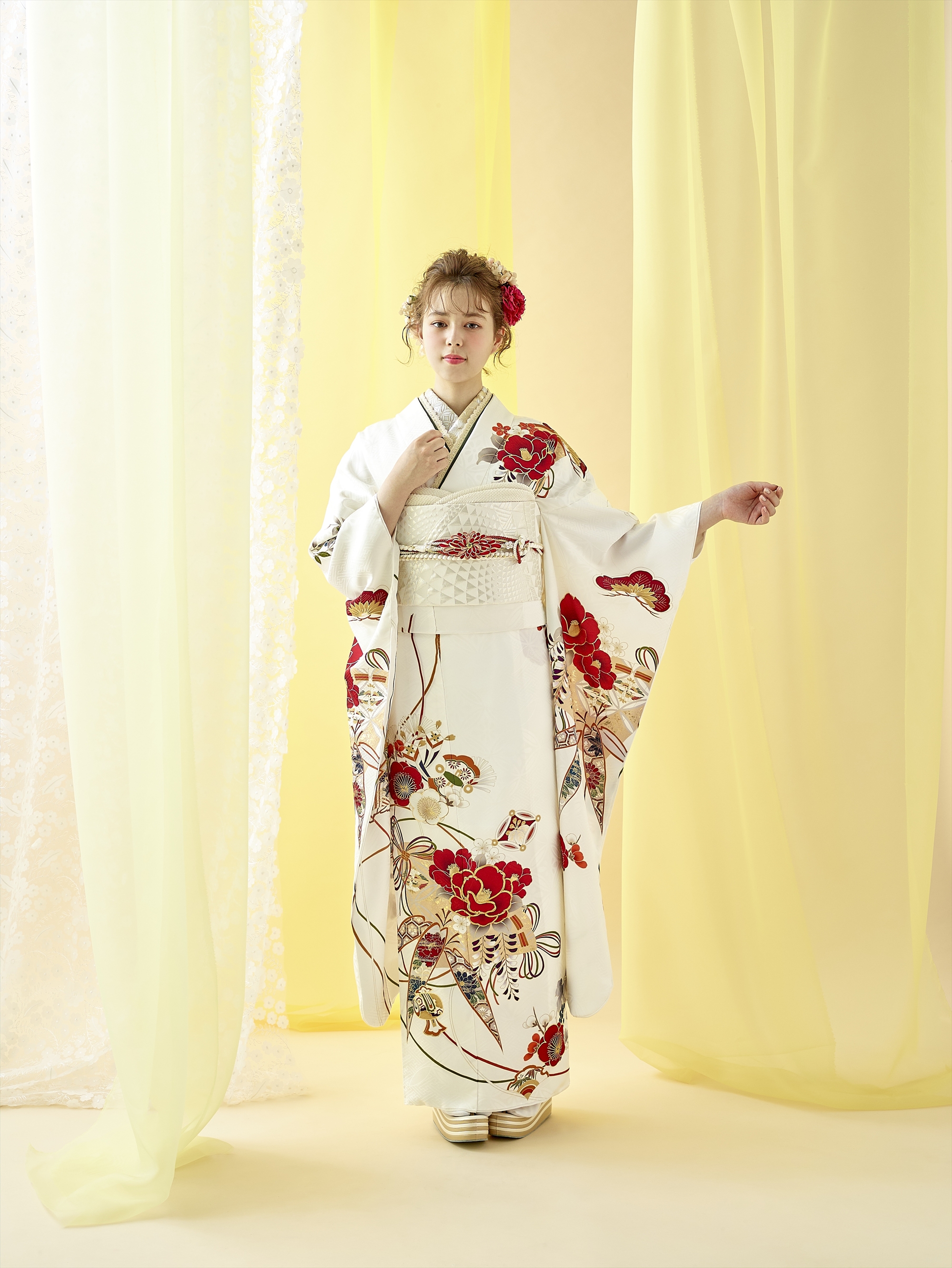 淡い黄色のカーテンを背に、赤と金の花柄が入った白い着物を着た若い女性が立っている。明るい髪をアップにし、赤い花飾りを添え、白い草履を履いている。右腕を上げ、着物の袖を見せている。.