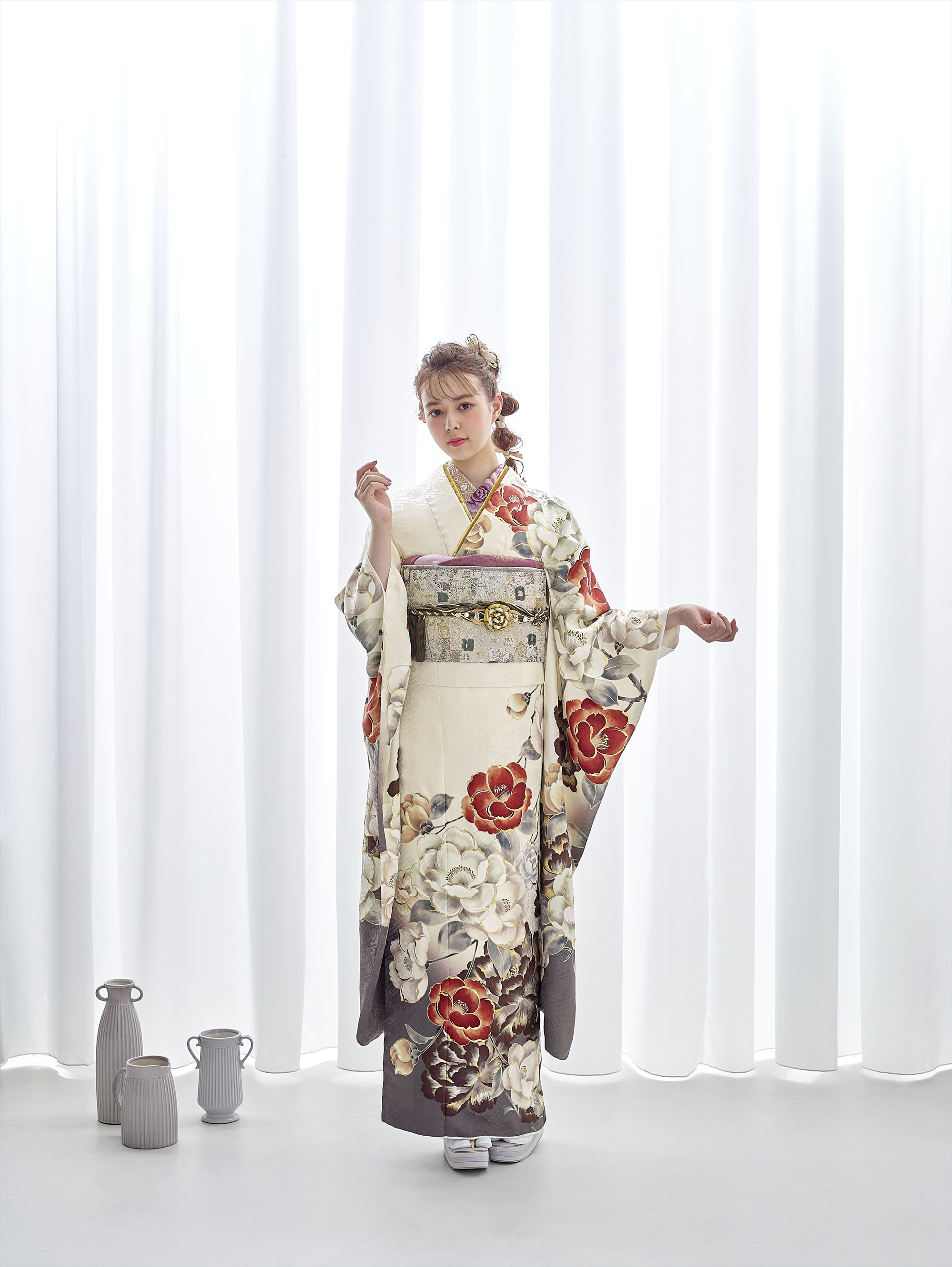 クリーム色の着物を着た女性が、白い幕の前に立っています。着物には紅白の大きな花模様が描かれています。華やかな髪飾りと白い草履を身につけています。彼女の左側の床には、陶器の水差しと花瓶が並べられています。.