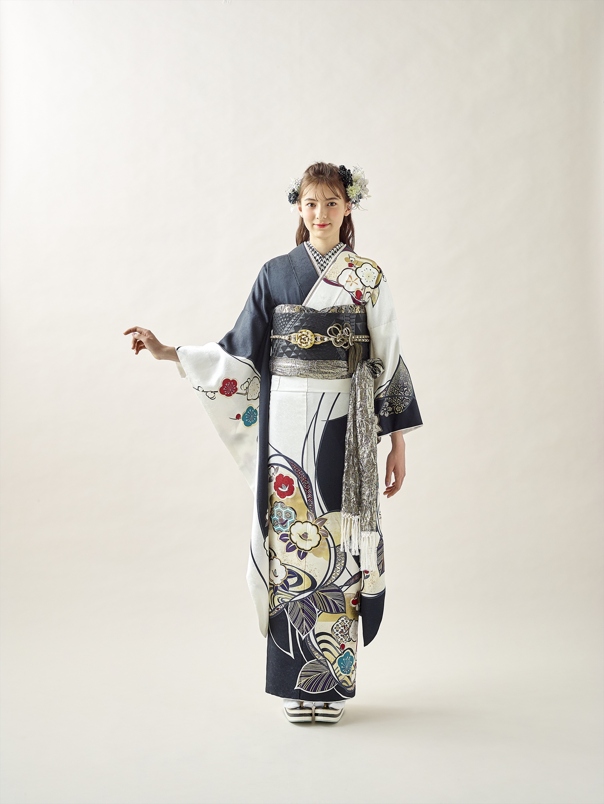 スタジオには、花柄の伝統的な白黒の着物と華やかな帯を締めた若い女性が立っています。髪には花を飾り、明るい背景に片腕を少し横に伸ばしています。.