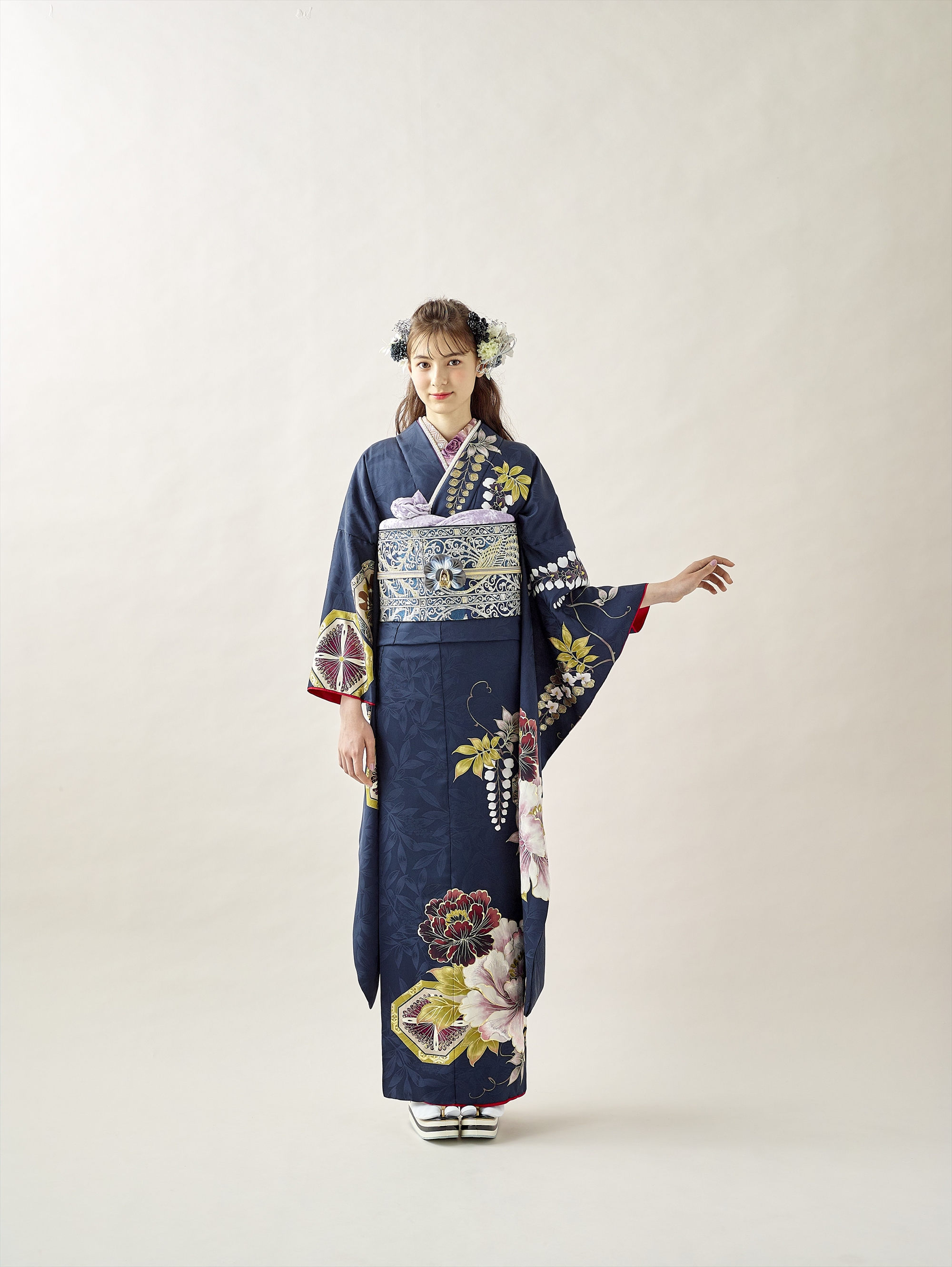 若い女性が、無地の背景に立っています。花柄の濃紺の着物に銀色の帯、白い足袋に草履を履いています。髪には花飾りが付けられ、片腕を伸ばし、もう片方の腕を体の横に置いて、正面を見つめています。.