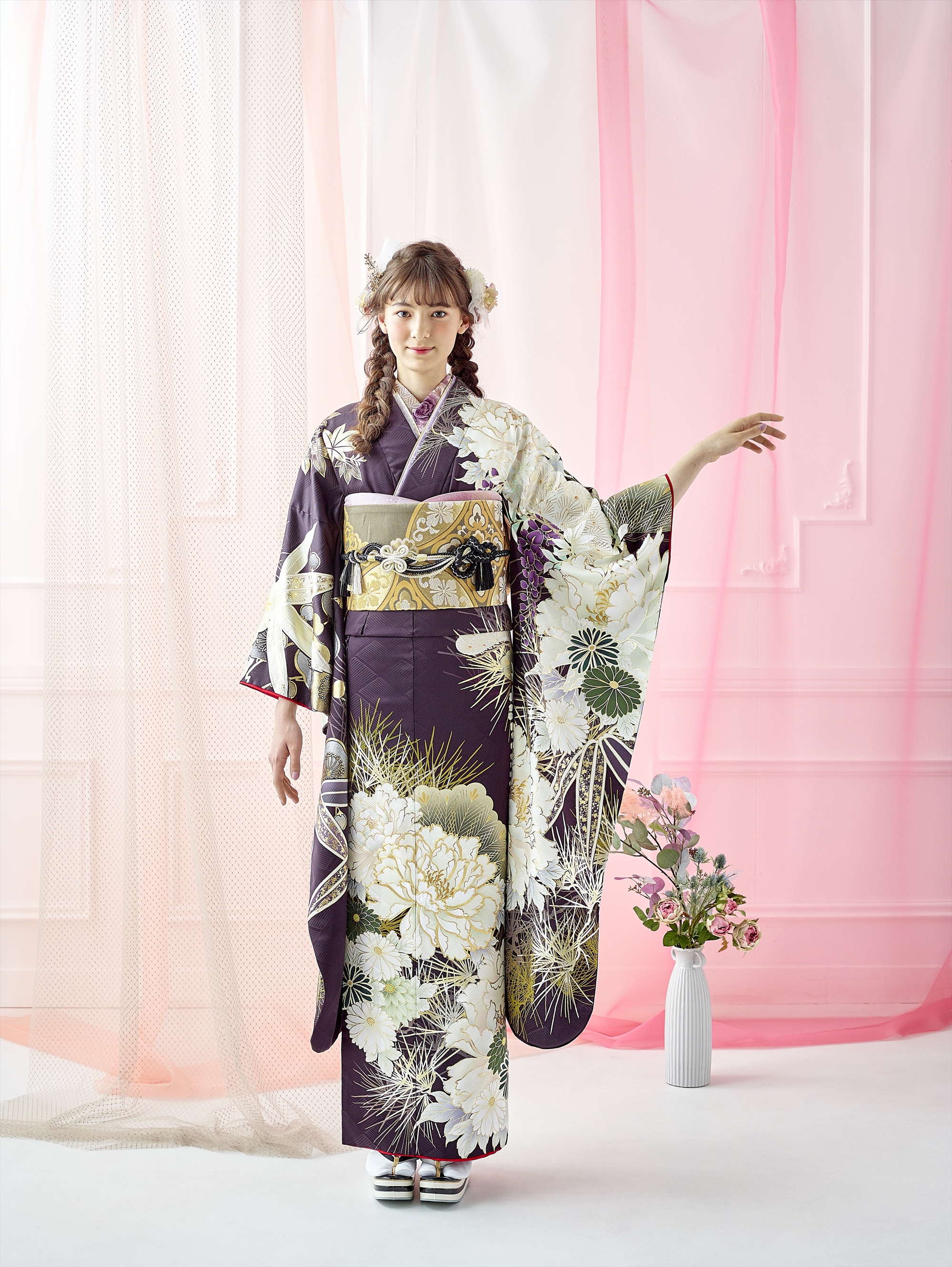 若い女性が、白と金の模様が入った濃い色の花柄の着物を着て、室内に立っています。片腕を上げてポーズをとっています。髪には花が飾られています。彼女の後ろには、白とピンクの透け感のあるカーテンがかかっており、床には背の高い花瓶が置かれています。.