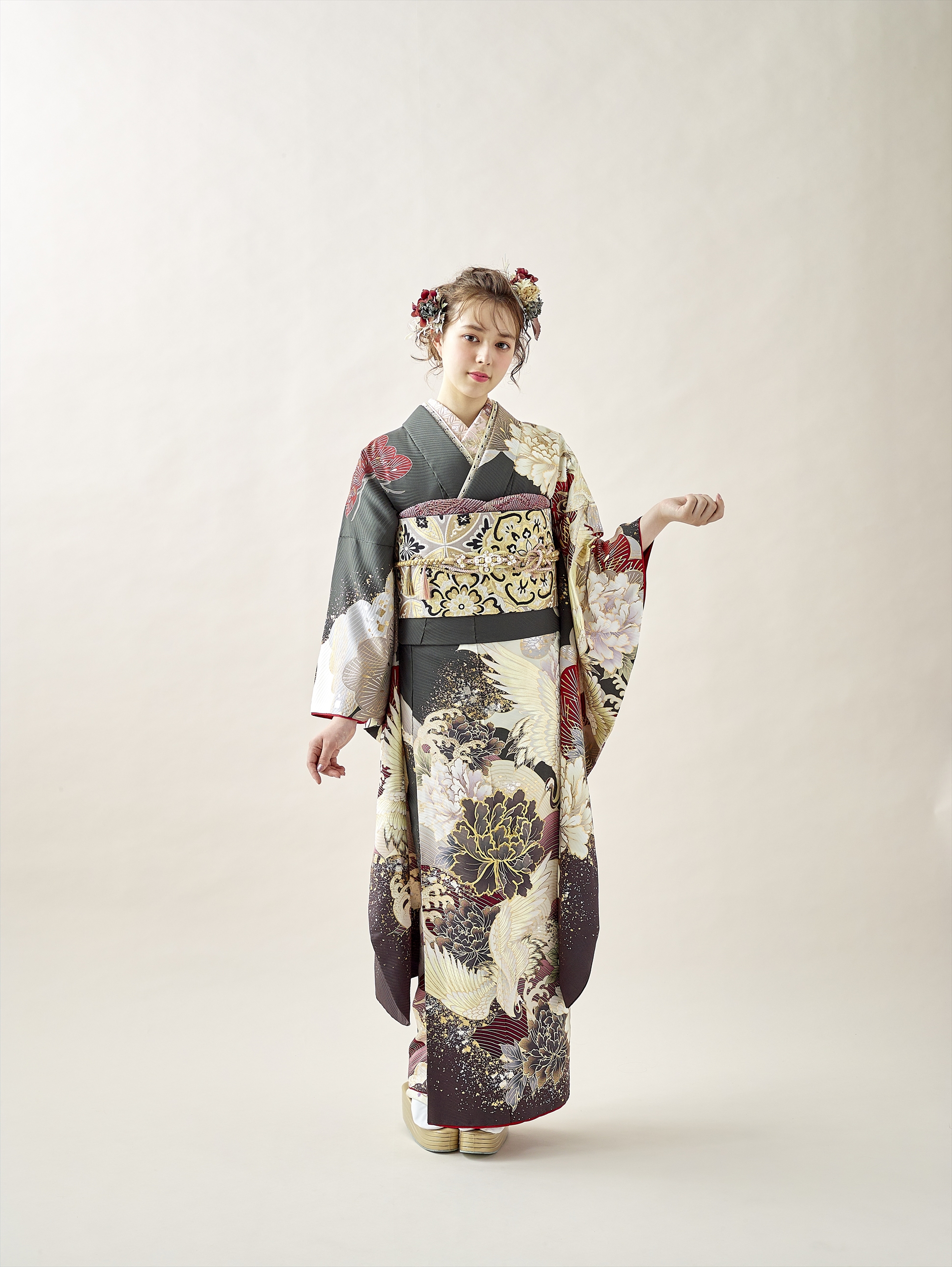花や鳥の模様があしらわれた華やかな着物を着た若い女性が、淡い色調の背景に立っています。赤と白の花飾りで髪を飾り、片腕を上げ、もう片方の腕を体の横に添えた優雅なポーズをとっています。.