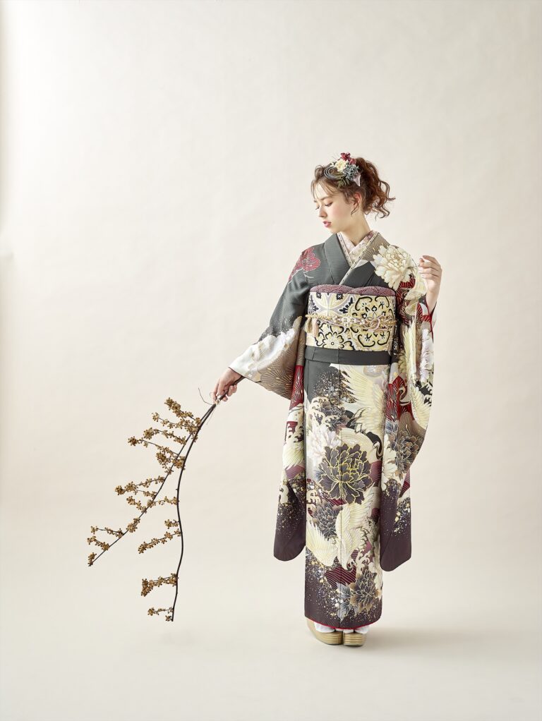 金、黒、象牙、赤の模様があしらわれた華やかな着物を着た女性が、小さな花が咲いた枝を持って立っています。彼女は花で飾られた豪華なアップヘアで、柔らかなベージュの背景に映える枝を見下ろしています。.