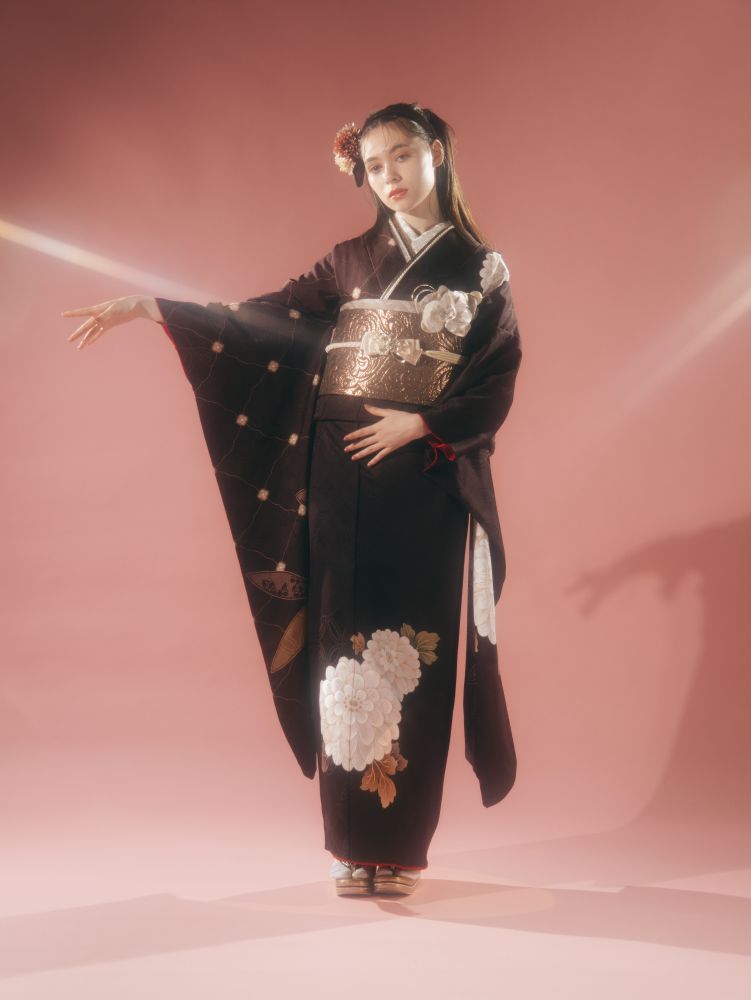 淡いピンクの背景に、白い花柄と金彩の細工が施された黒い着物をまとった人物が優雅に立っています。髪には花飾りが付けられ、片方の腕は優雅に伸ばされ、着物の袖が露わになっています。もう片方の腕は腰のあたりに添えられています。.