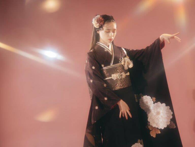 淡いピンクの背景に、大きな白い花柄があしらわれた伝統的な黒の着物をまとった女性が優雅にポーズをとっています。髪はセットされ、花飾りがアクセントになっています。彼女はうつむき、優雅な視線を向けています。柔らかな光の反射が、夢のような雰囲気を醸し出しています。.