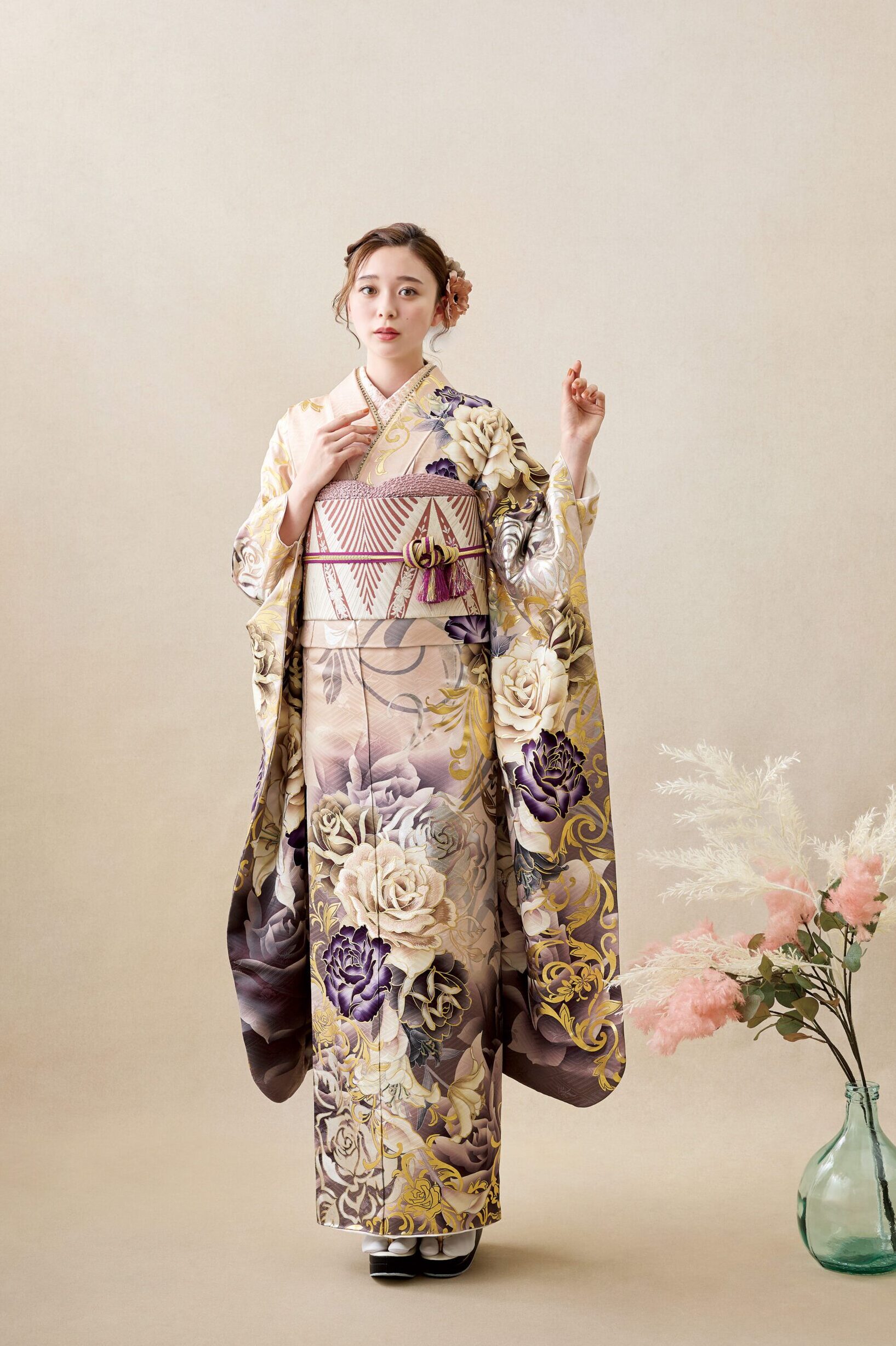 ベージュの背景に、金、紫、クリーム色の花柄があしらわれた着物4053（複製）を着た女性がポーズをとっている。髪には花を挿し、袖を持ち、傍らにはピンクと白の花が入ったガラスの花瓶が置かれている。.