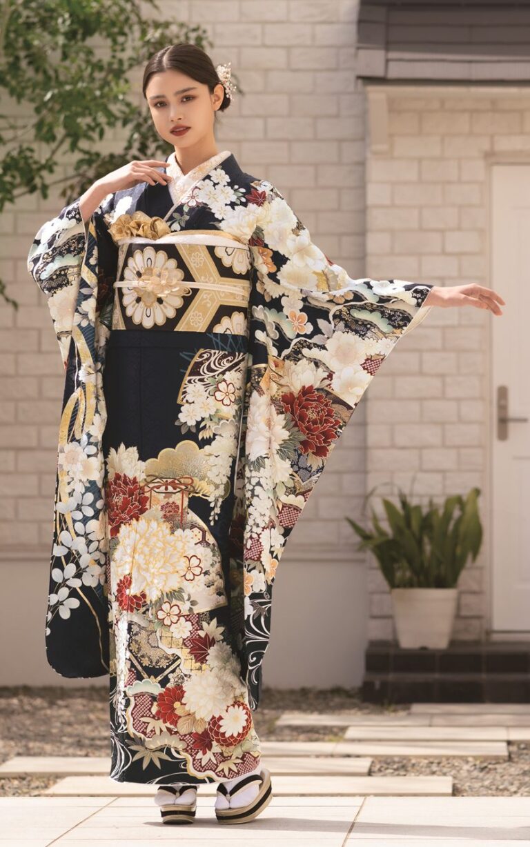 モダンな白いレンガ造りの家の外に、濃い花柄の袖の広い着物を着て金の帯を締めた女性が立っている。両手を広げて優雅にポーズをとっている。明るく日当たりの良い場所で、近くには小さな木と植物がある。.