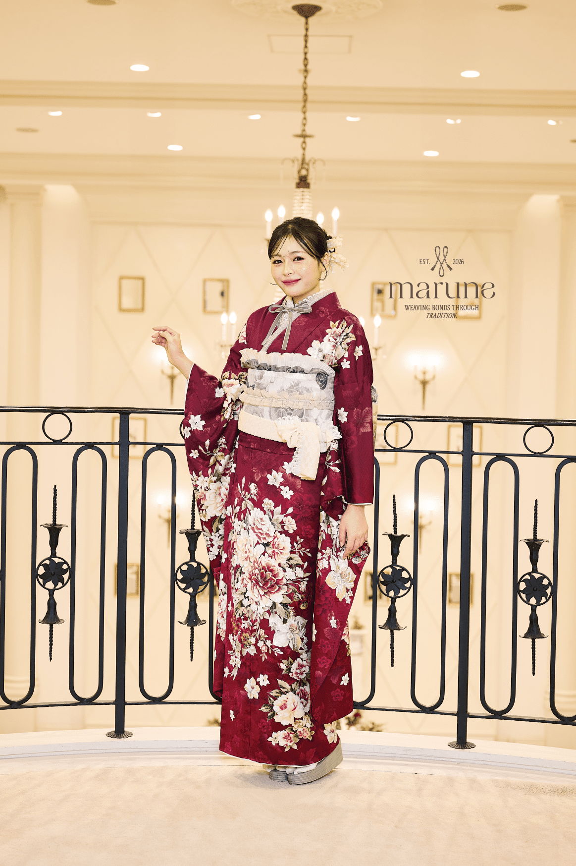 室内のバルコニーで優雅にポーズをとる女性が、薄手の帯を締めた深紅の花柄の着物m-601（複製）を着て微笑んでいる。エレガントな照明、シャンデリア、クリーム色の壁、丸根の看板が洗練されたシーンを完成させている。.